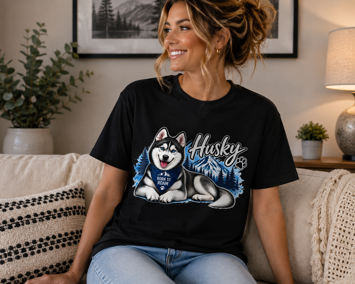 A woman sitting on a couch wearing a black graphic t-shirt with a cartoon husky and the text 'Husky PUPS DOUGH' on the front, in a cozy living room setting.